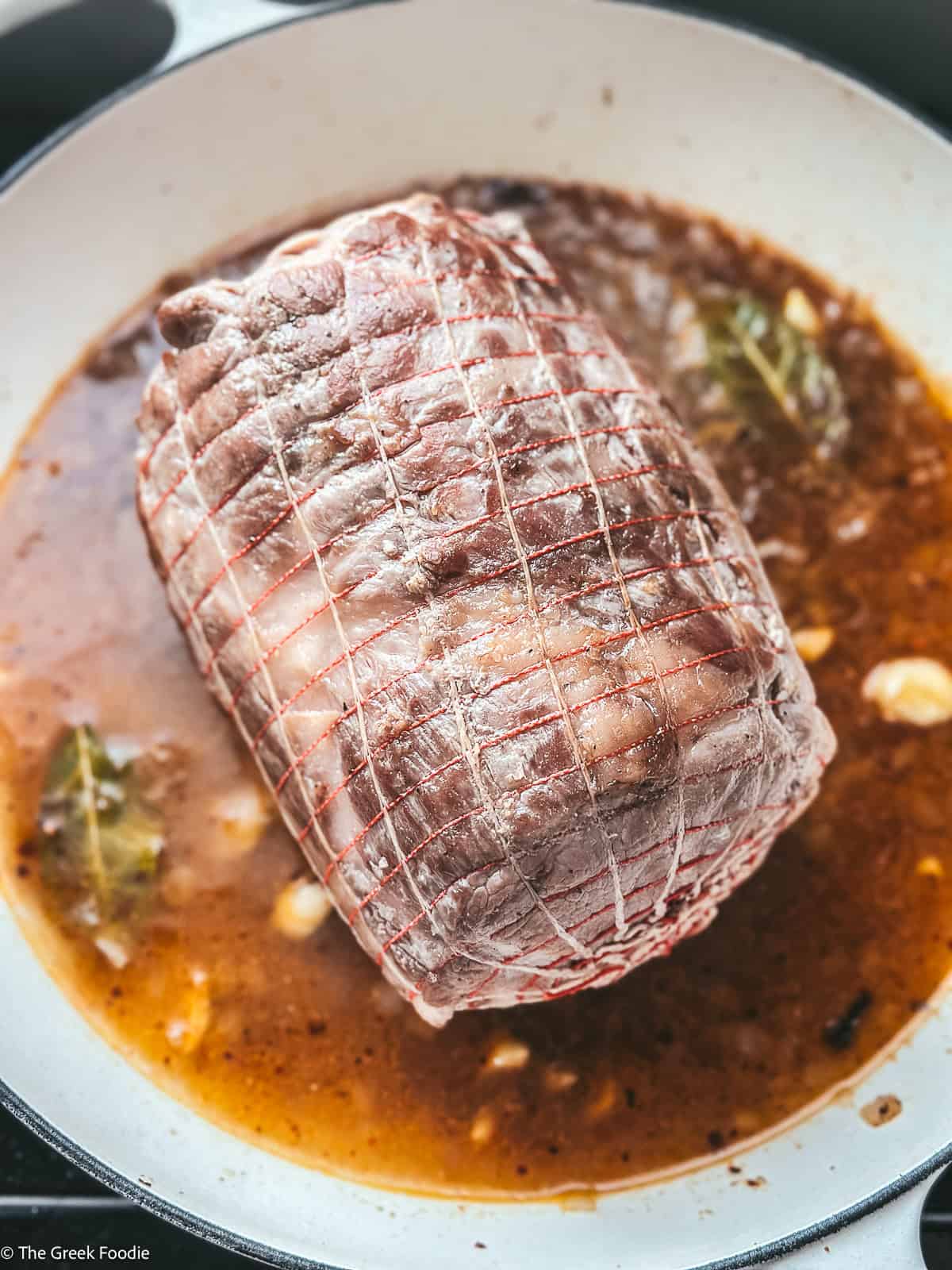 A pork shoulder wrapped in a net with broth with onion, bay leaves and seasonings cooking in a white pot.