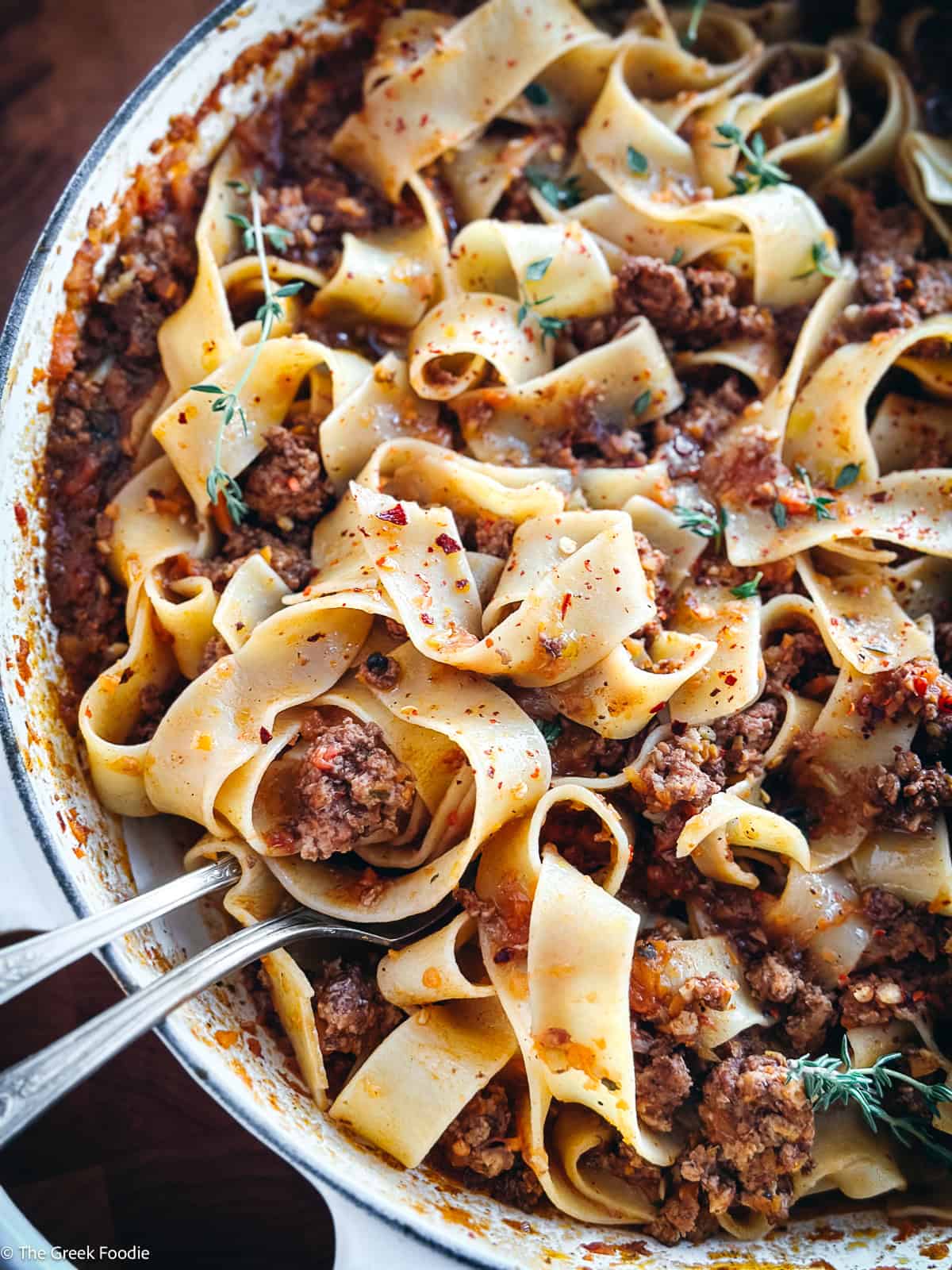 A white dutch oven with chicken bolognese with pappardelle pasta and two serving utensils on a table.