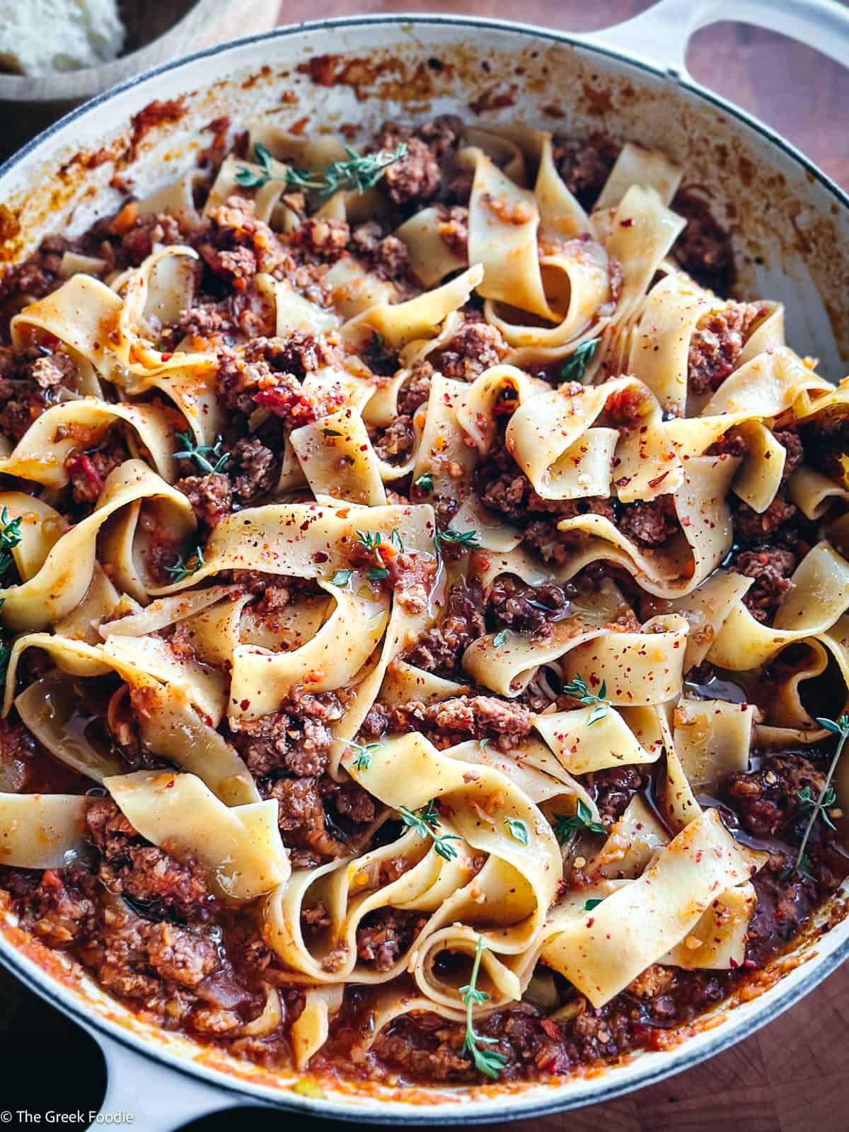 A white dutch oven with chicken bolognese with papardele pasta and two serving utensils on a table.