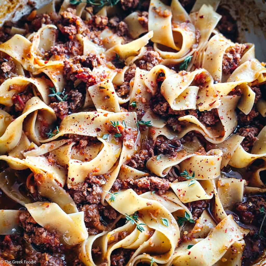 A white dutch oven with chicken bolognese with papardele pasta and two serving utensils on a table.