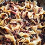 A white dutch oven with chicken bolognese with papardele pasta and two serving utensils on a table.