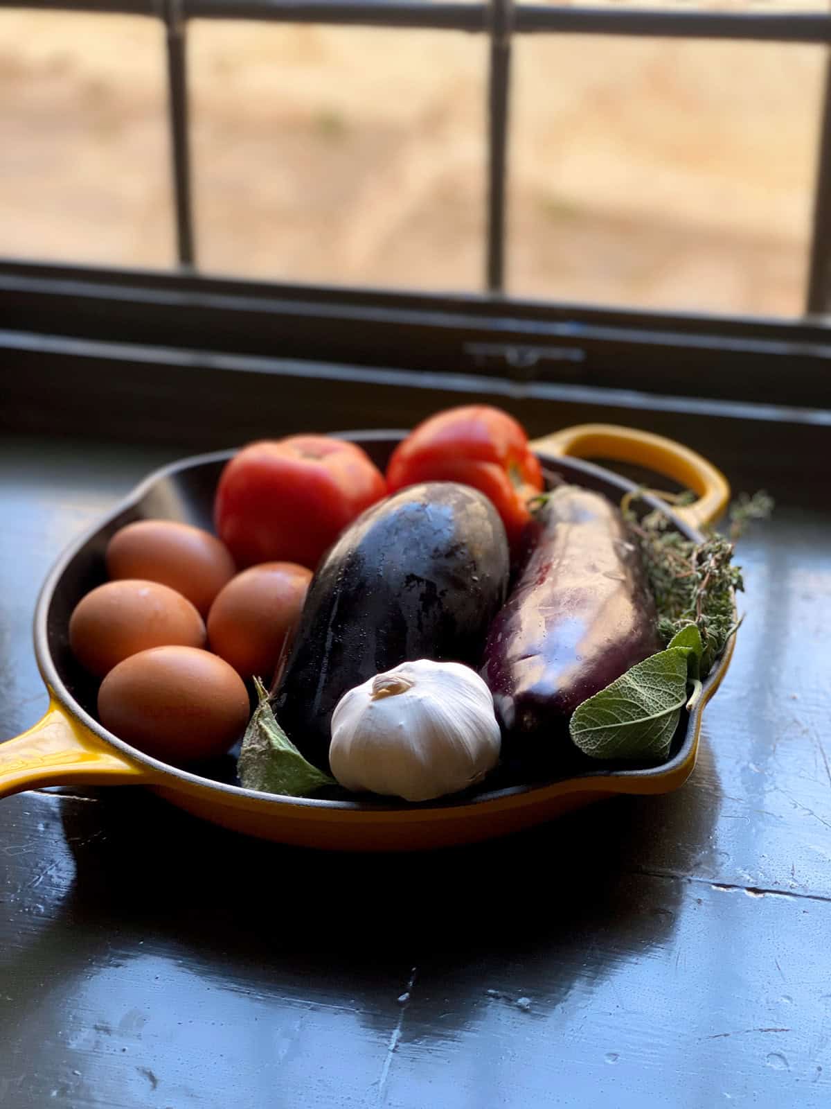 Eggplants, eggs, tomatoes in a yellow skillet by a window.