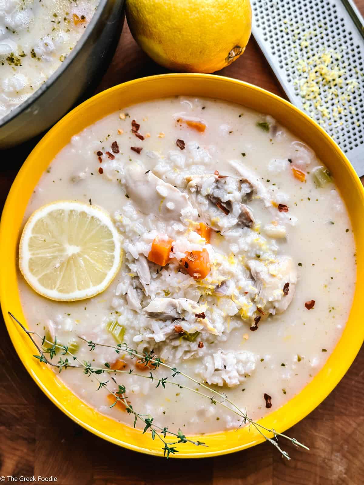 A yellow deep plate with greek chicken soup with a lemon slice on a table.