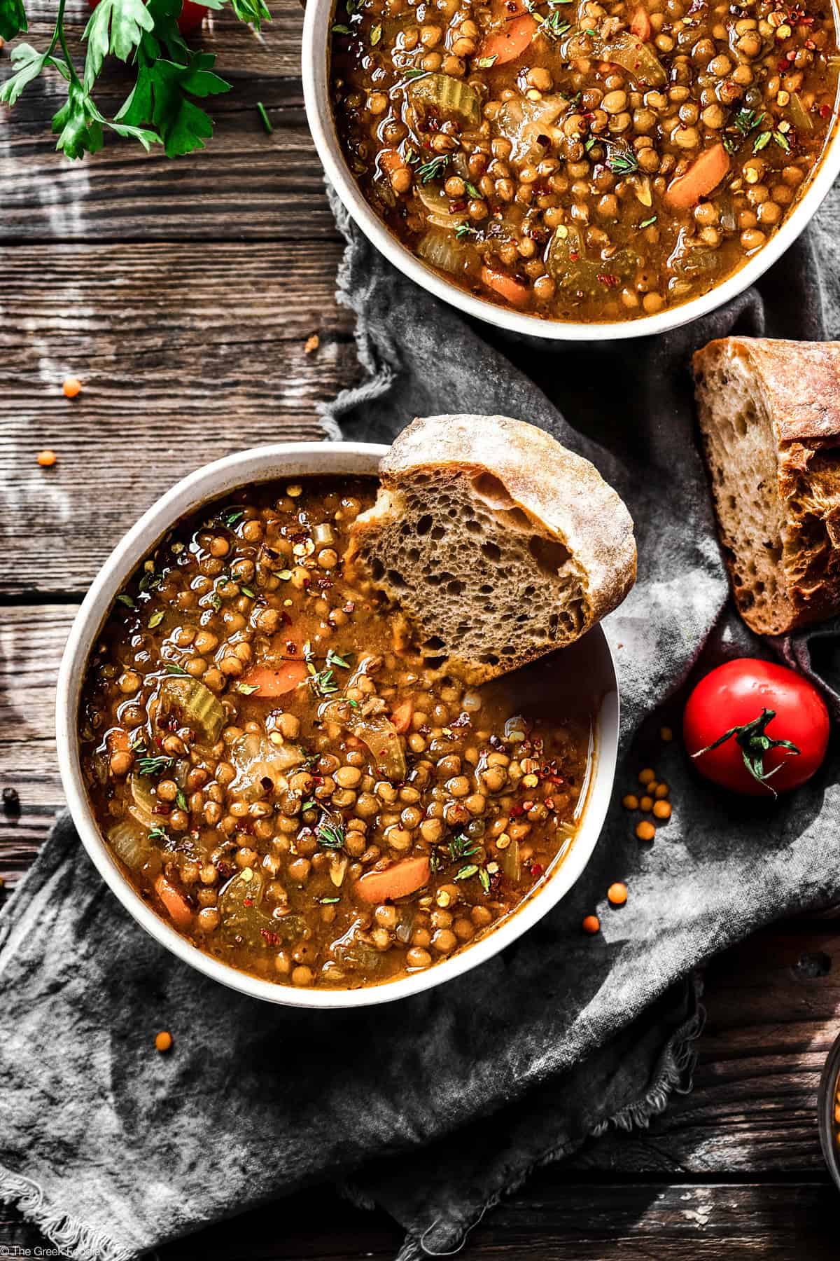 Two bowls with Greek lentil soup, bread and a tomato on a table.