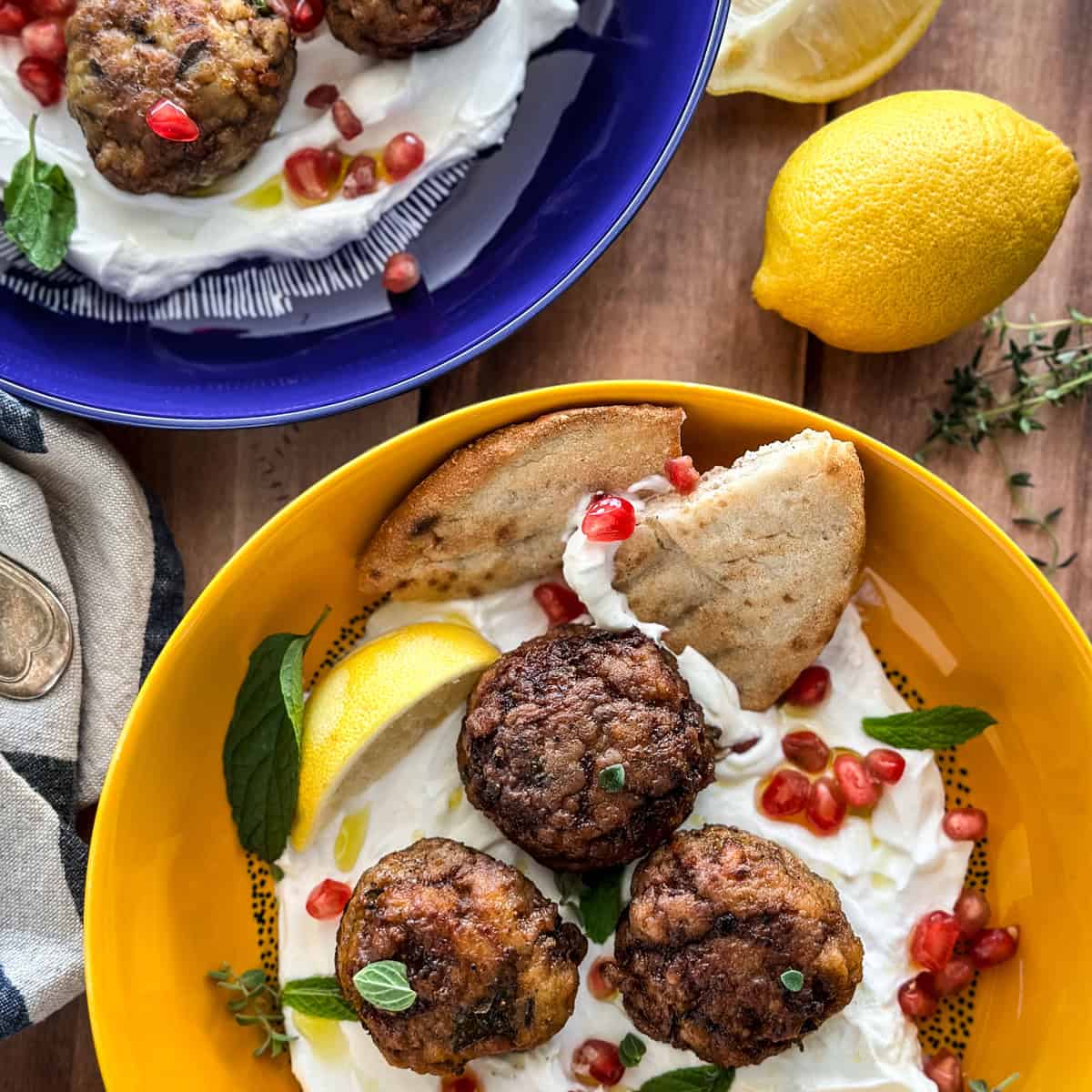 Two plates with lamb meatballs over yogurt with pomegranate seeds, pita and lemon wedges.