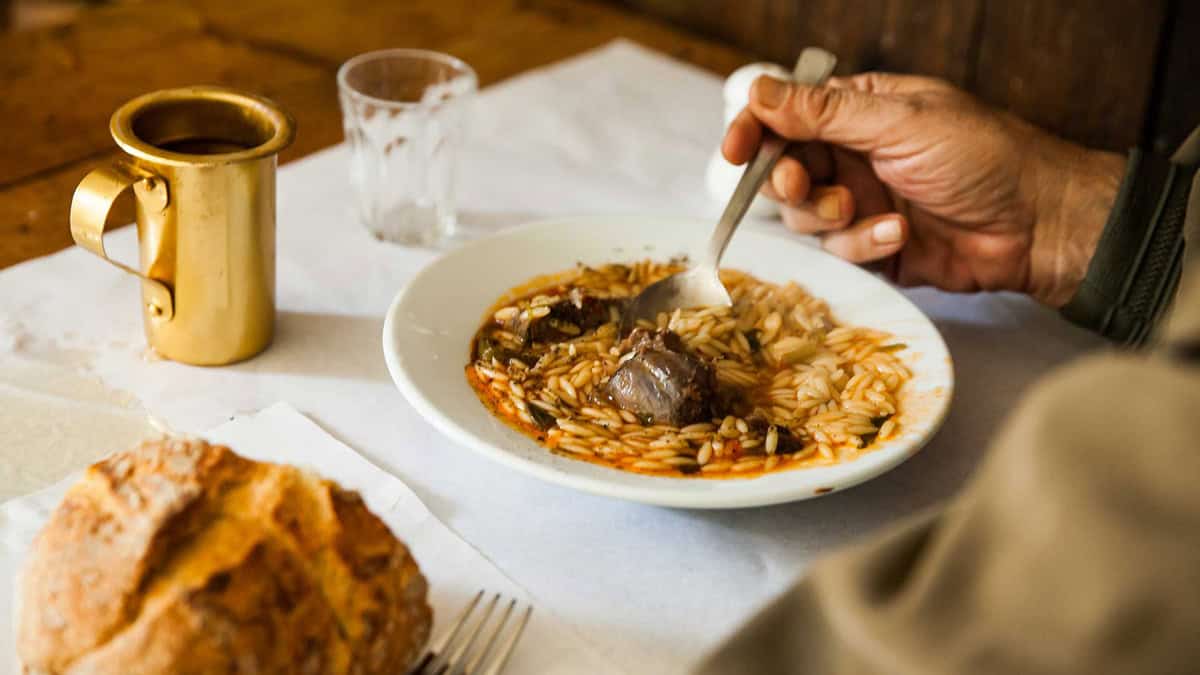 A hand holding a spoon, scooping orzo from a plate with youvetsi, on the table a metallic jug for wine.