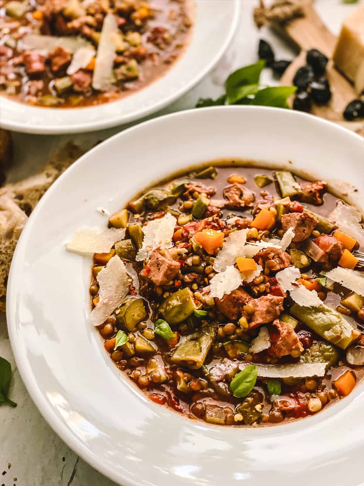 A bowl with lentil sausage soup, partial view of another bowl, some bread, and a tray with olives and parmesan cheese.