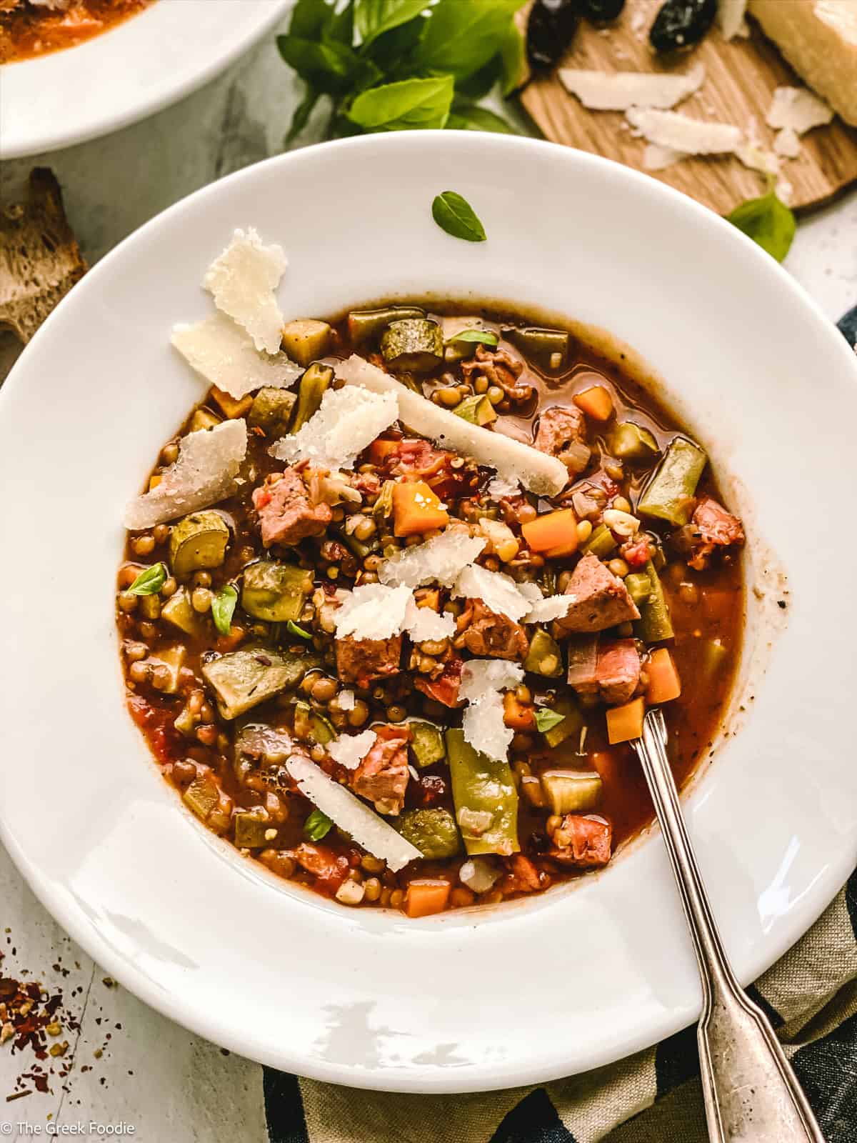 A bowl with lentil sausage soup, partial view of another bowl, some bread, and a tray with olives and parmesan cheese