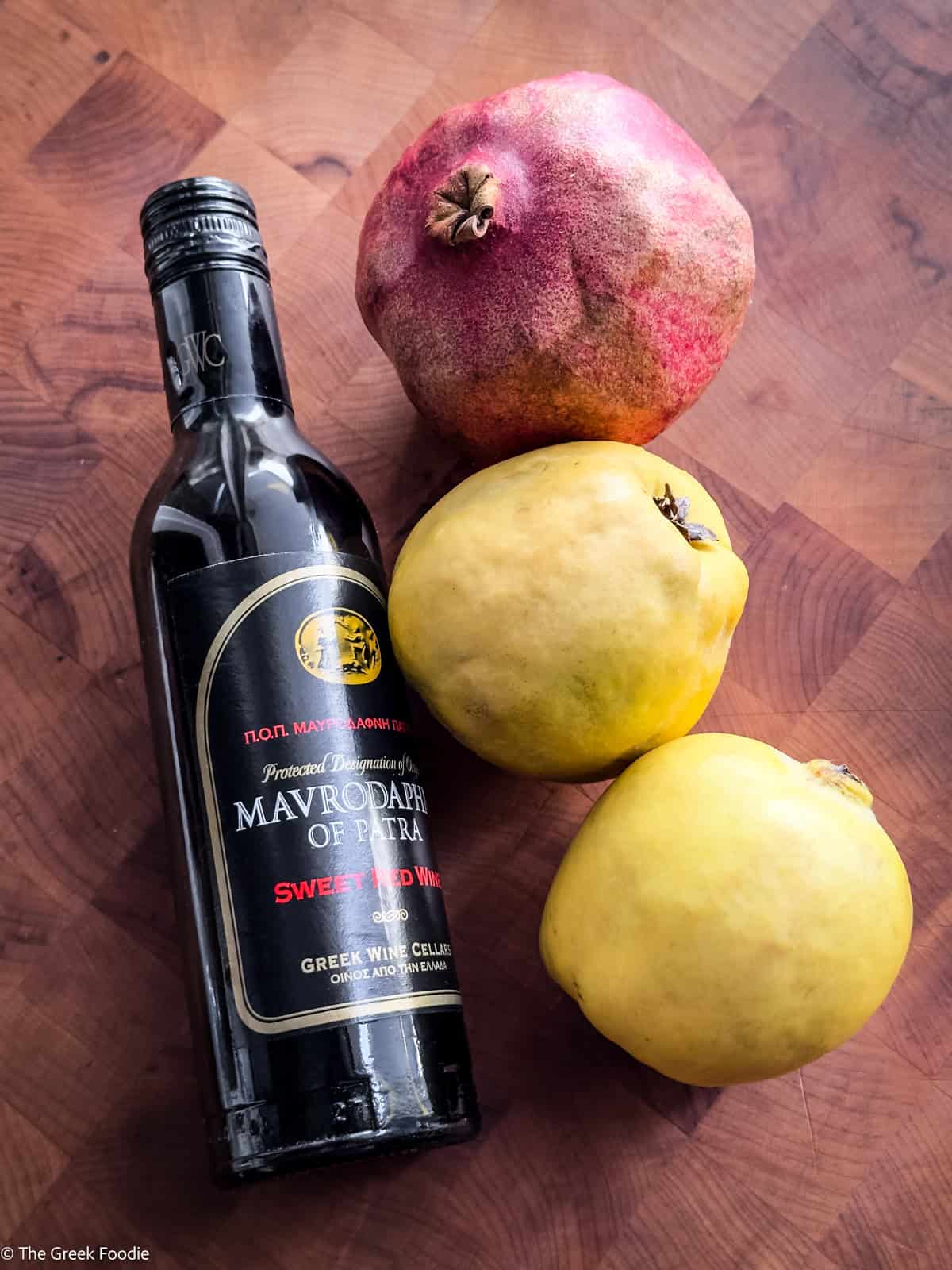 A bottle of mavrodaphne wine next to two quinces and a pomegranate on a wooden surface.