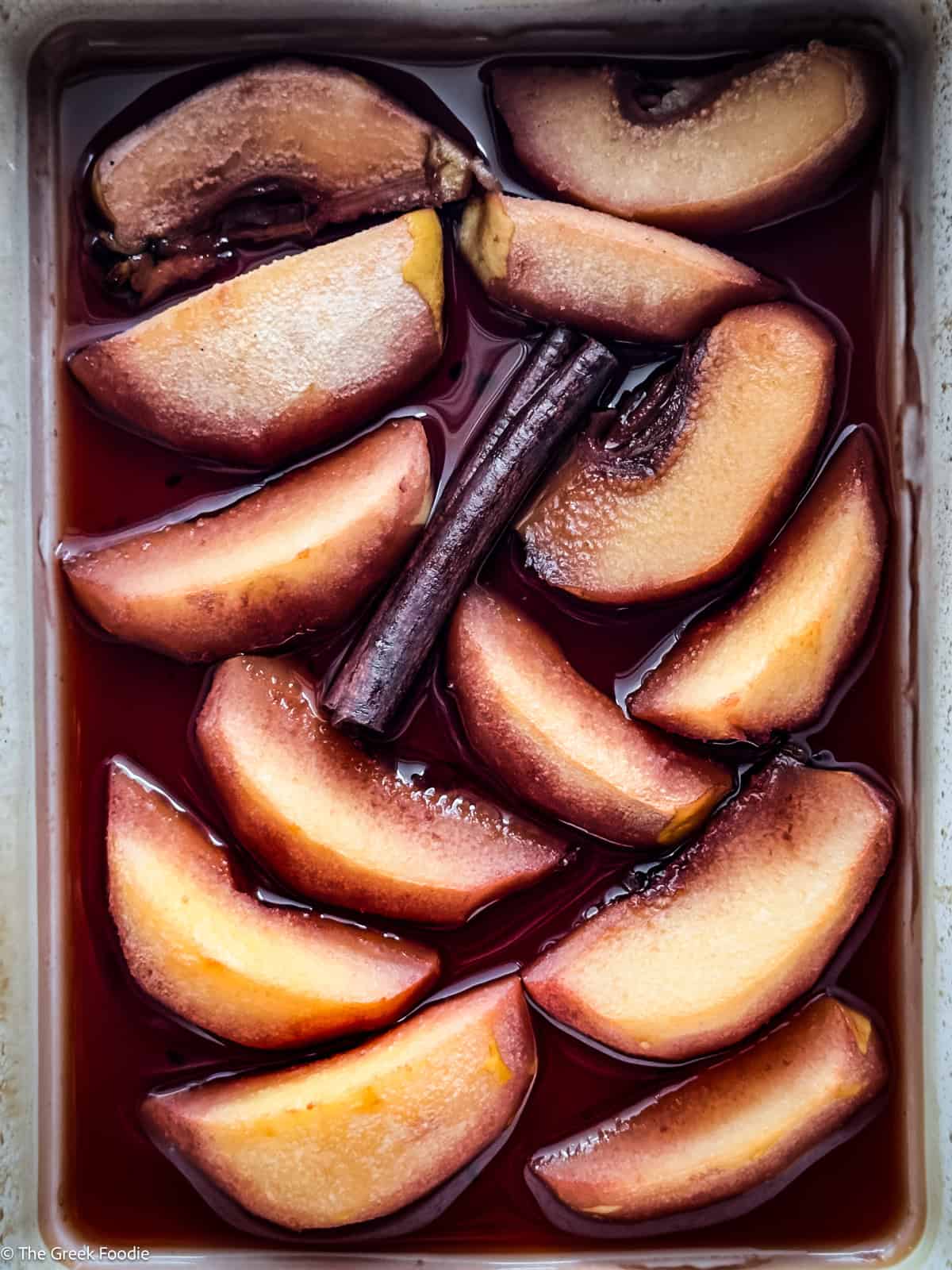 Roasted sliced quince with a cinnamon stick and red wine in a white pan.