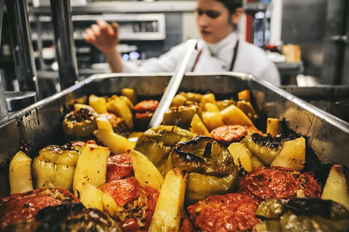 At the front a pan with stuffed tomatoes, behind it blurry a lady chef.