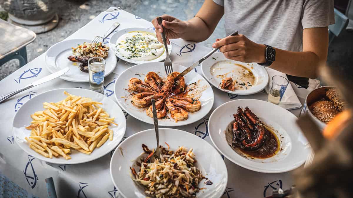 A table with Greek food and the hands of a woman serving food to her plate.