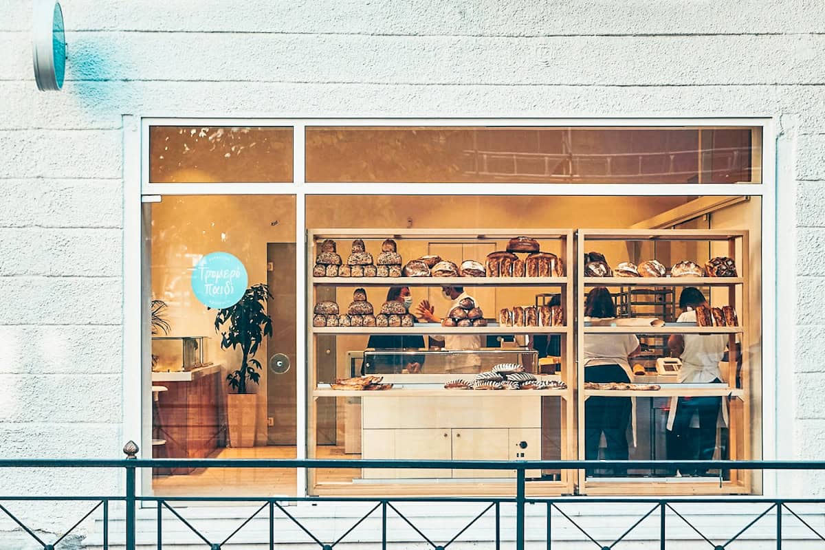 The storefront of Tromero Paidi bakery in Athens is white with big windows with white trim, showing the shelves full of bread loaves.