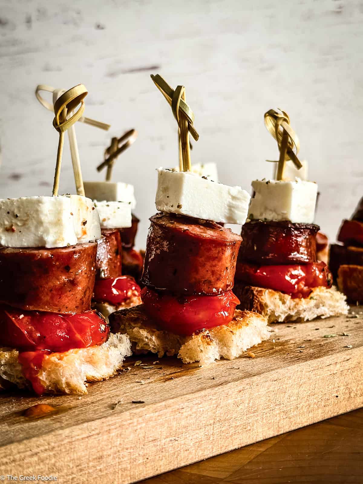 Greek sausage bites with tomato confit, feta cheese, and crusty bread served on wooden board.