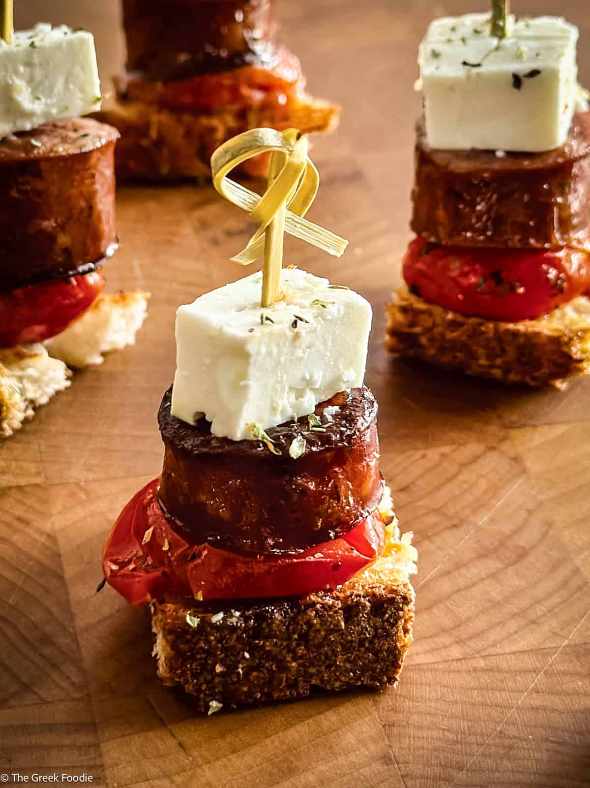Greek sausage crostini topped with tomato confit and feta cheese, secured with toothpicks.