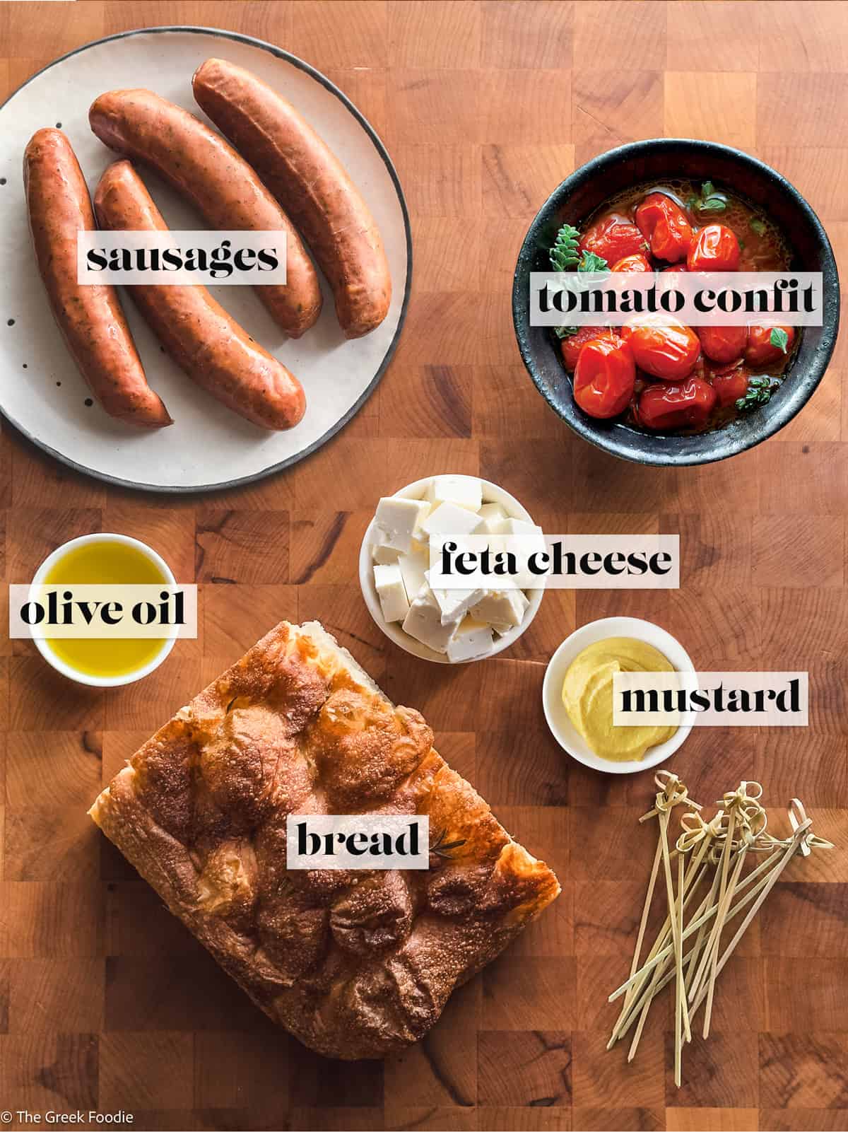 Ingredients for Greek sausage bites including sausages, tomato confit, feta, bread, olive oil, and mustard.