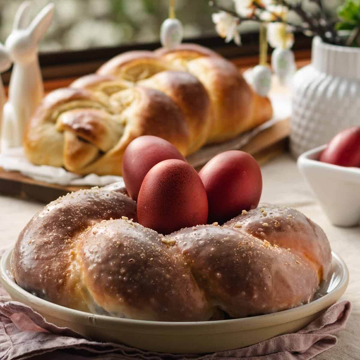 A Tsoureki bread with red eggs in the middle on a table, at the back more Tsoureki bread and flowers, on a table.