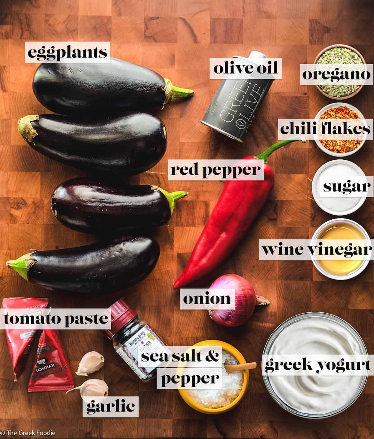 Four eggplants, a red pepper, garlic, onion and tomato paste tubes, sea salt and pepper, and bowls of seasonings, and a bowl of yogurt on a wooden surface.