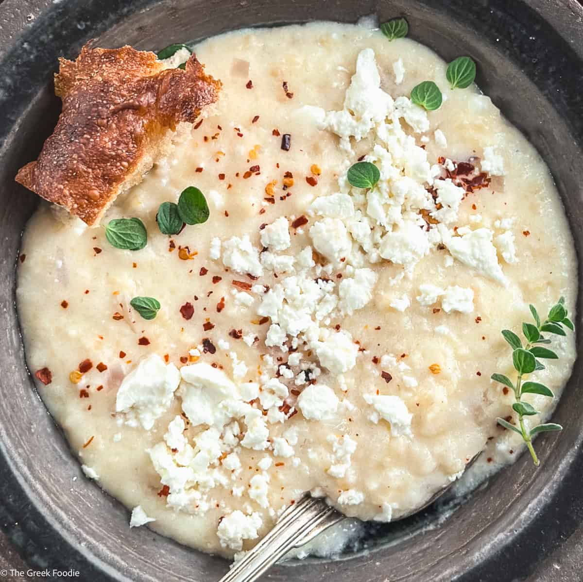 Creamy traditional Greek trahanas soup made with butter and feta, topped with cracked black pepper and chili flakes, served with crusty bread.