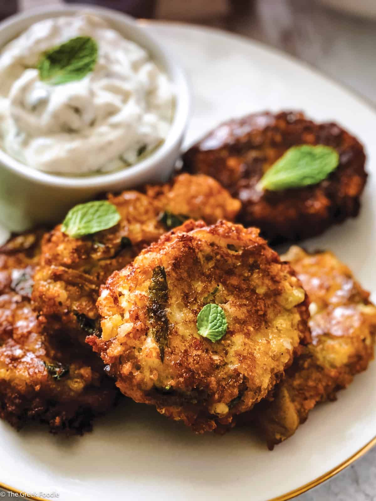 Four eggplant fritters on a plate with a container with yogurt dip, some fresh basil leaves and a cloth napkin at the back.