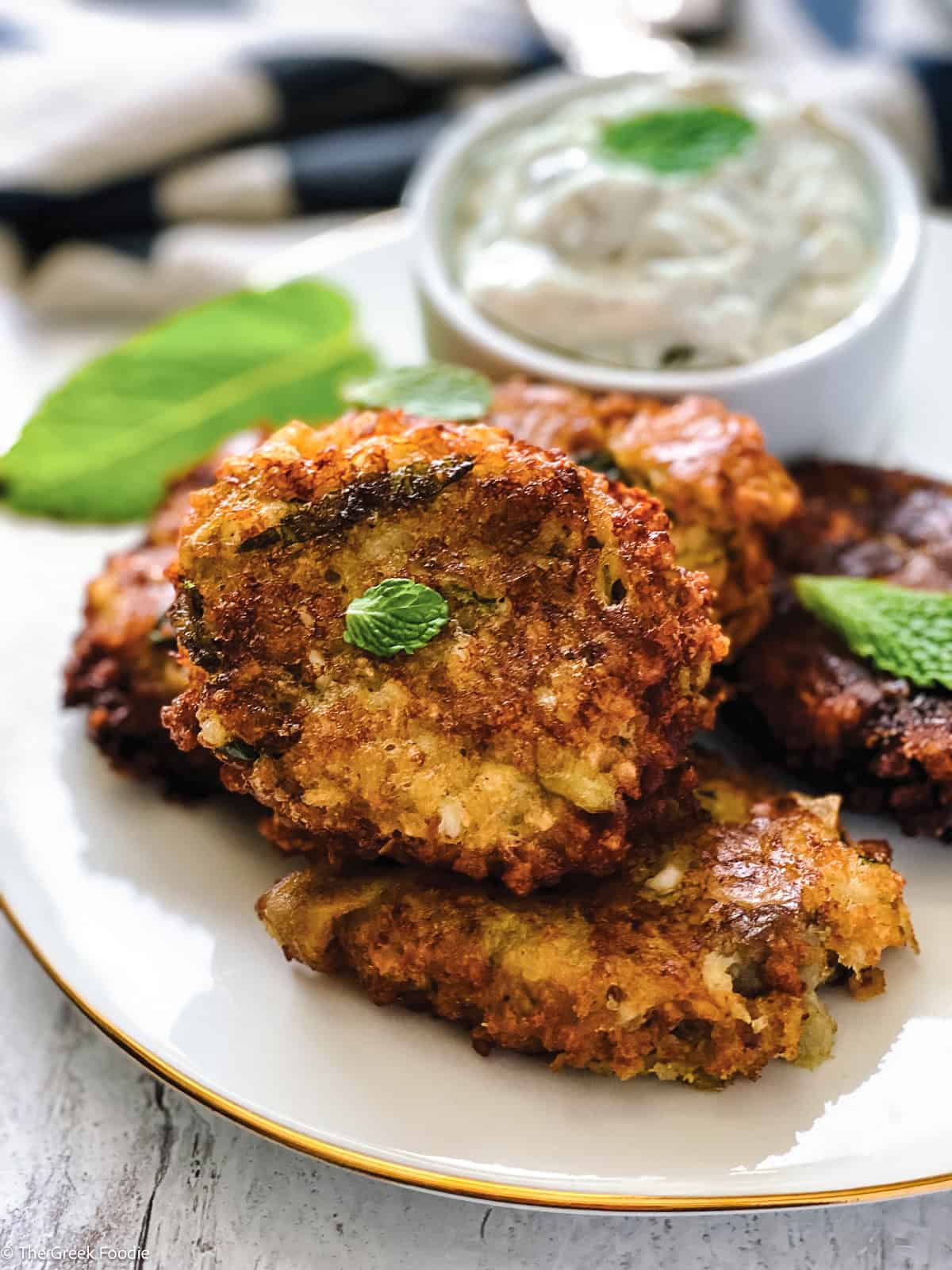 Four eggplant fritters on a plate with a container with yogurt dip, some fresh basil leaves and a cloth napkin at the back.