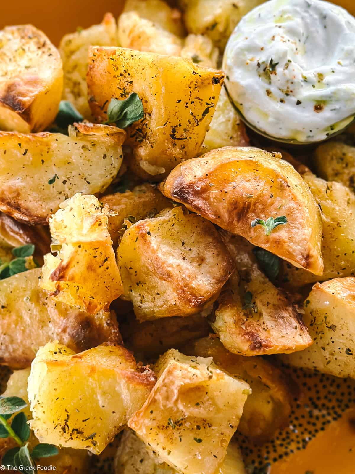 Close-up of golden roasted potato bites seasoned with oregano, served with a small cup of creamy tzatziki for dipping.