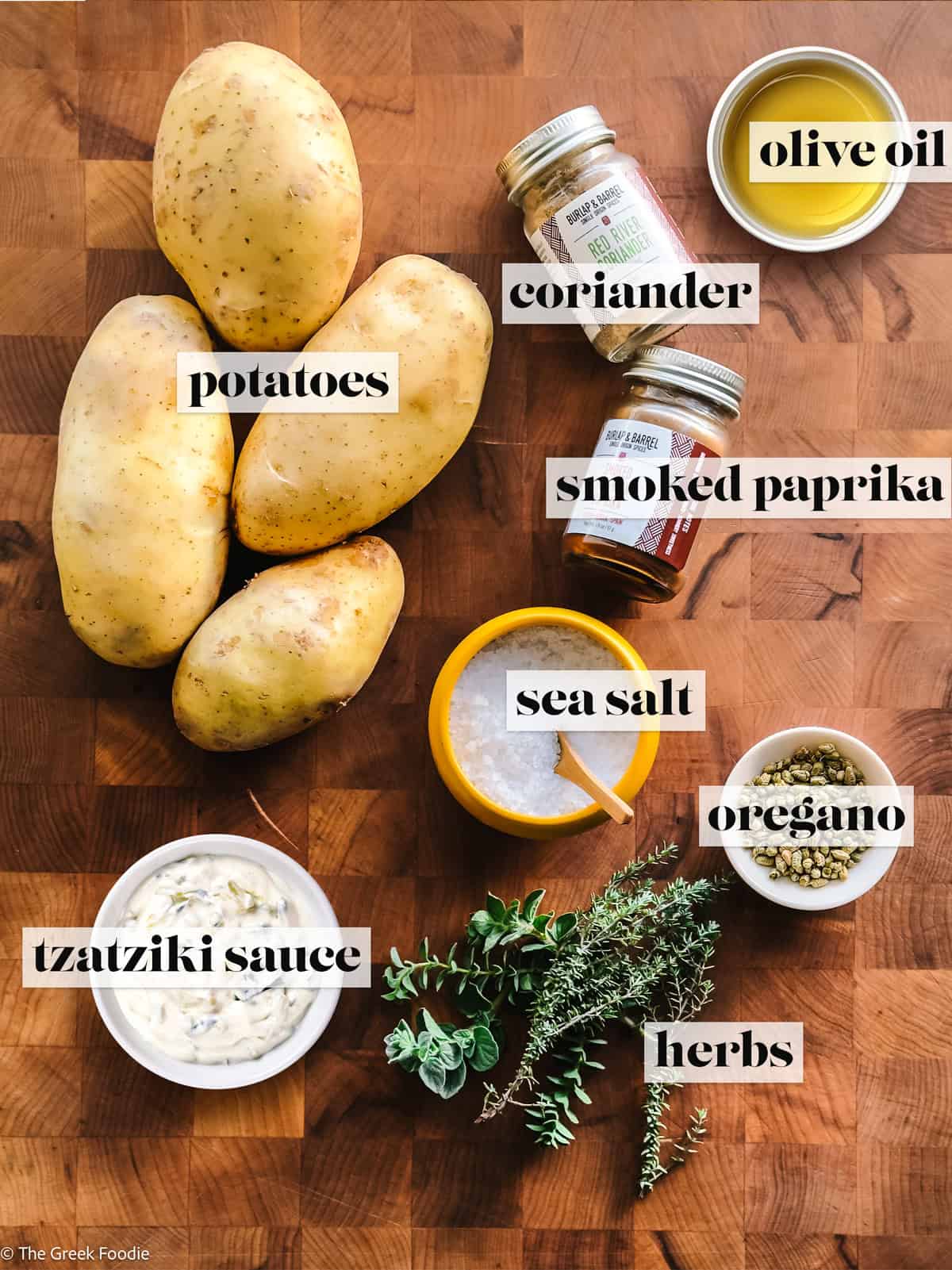 Ingredients for Greek-style potatoes including whole potatoes, olive oil, sea salt, oregano, coriander, smoked paprika, and fresh herbs on wooden board.