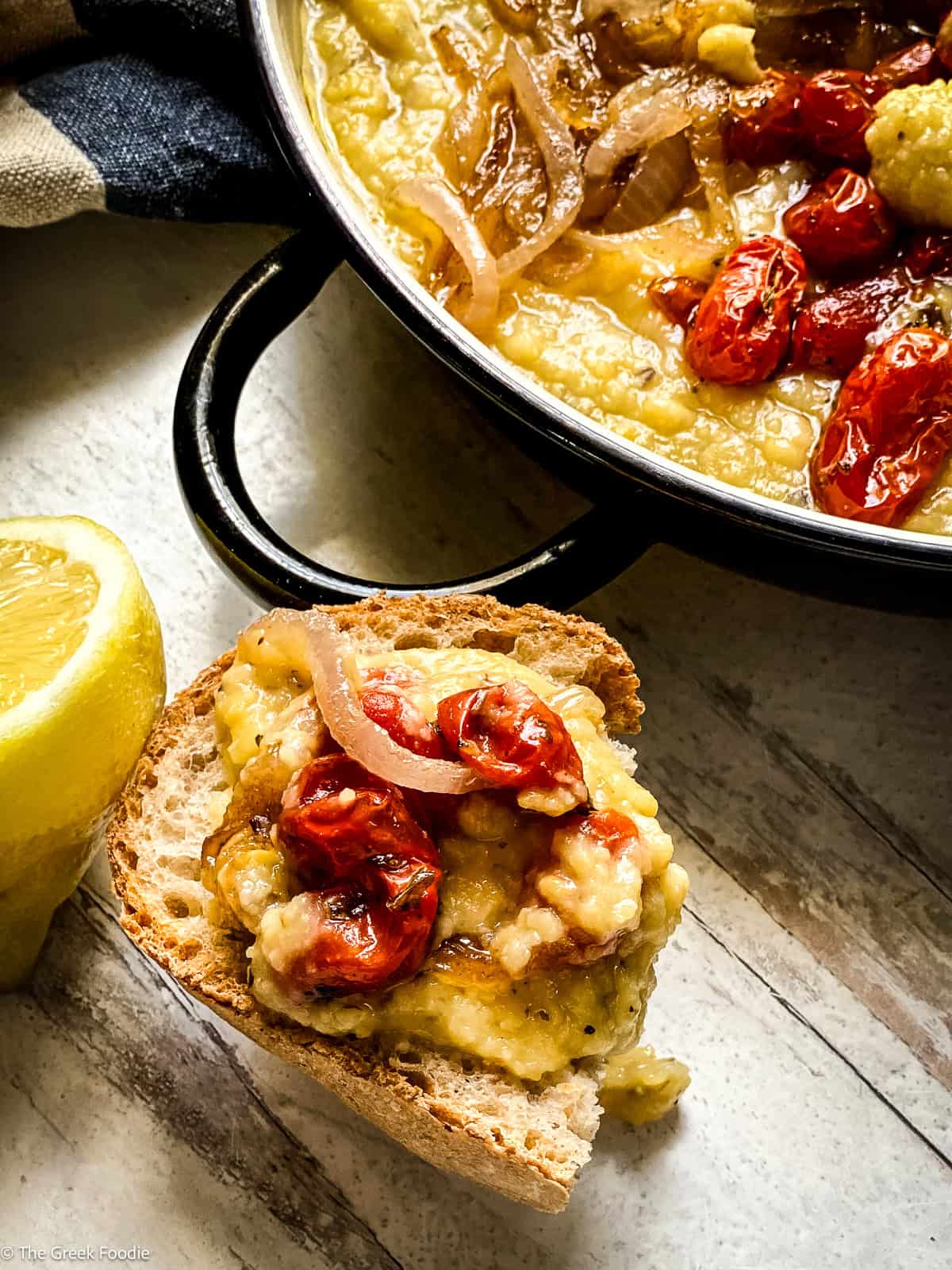 A piece of crusty bread topped with Greek fava dip, roasted cherry tomatoes, and caramelized onions with a lemon wedge on the side.