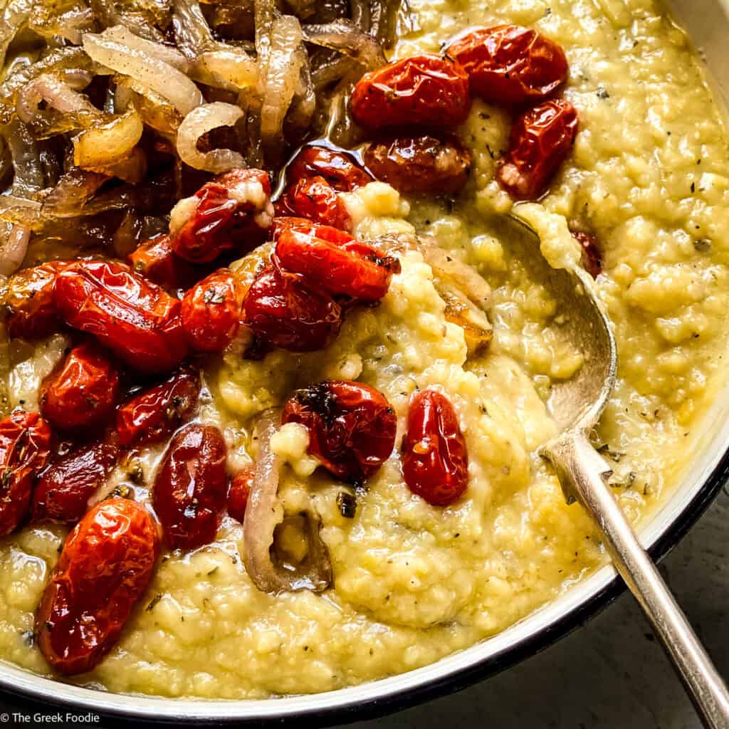 Close-up of Greek fava dip in a serving pan topped with burst roasted tomatoes and caramelized onions with a spoon.