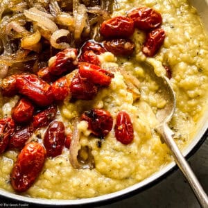 Close-up of Greek fava dip in a serving pan topped with burst roasted tomatoes and caramelized onions with a spoon.
