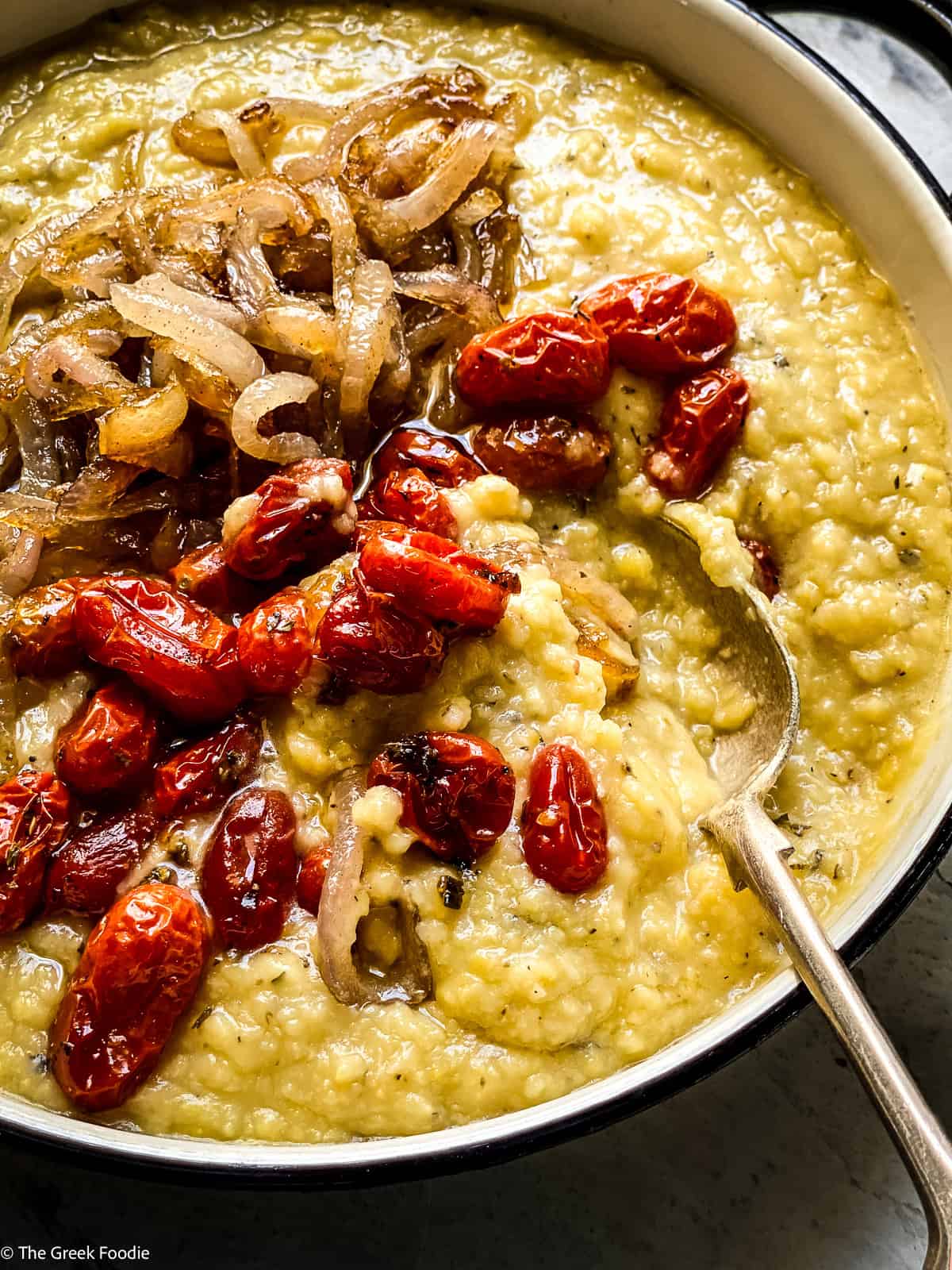 Close-up of Greek fava dip in a serving pan topped with burst roasted tomatoes and caramelized onions with a spoon.
