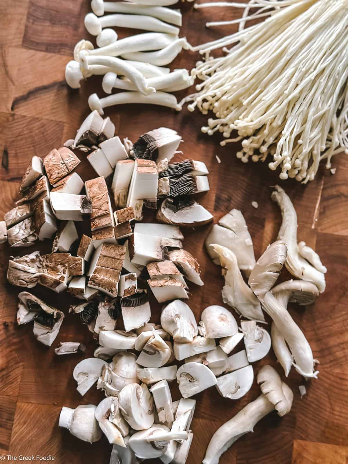 Five mushrooms prepped for Greek mushroom magiritsa — button, portobello, oyster, shimeji and enoki on a wooden board