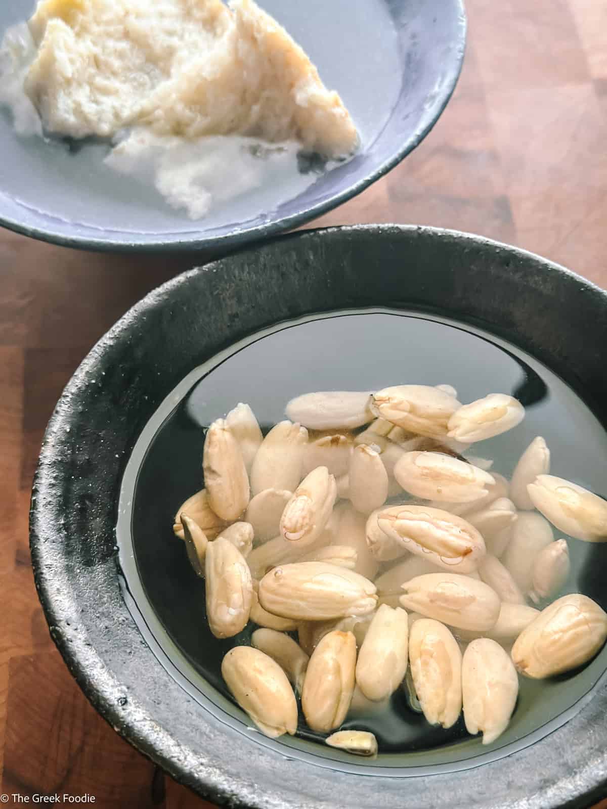 Blanched almonds soaking in water with soaked bread in the background, for making almond skordalia.