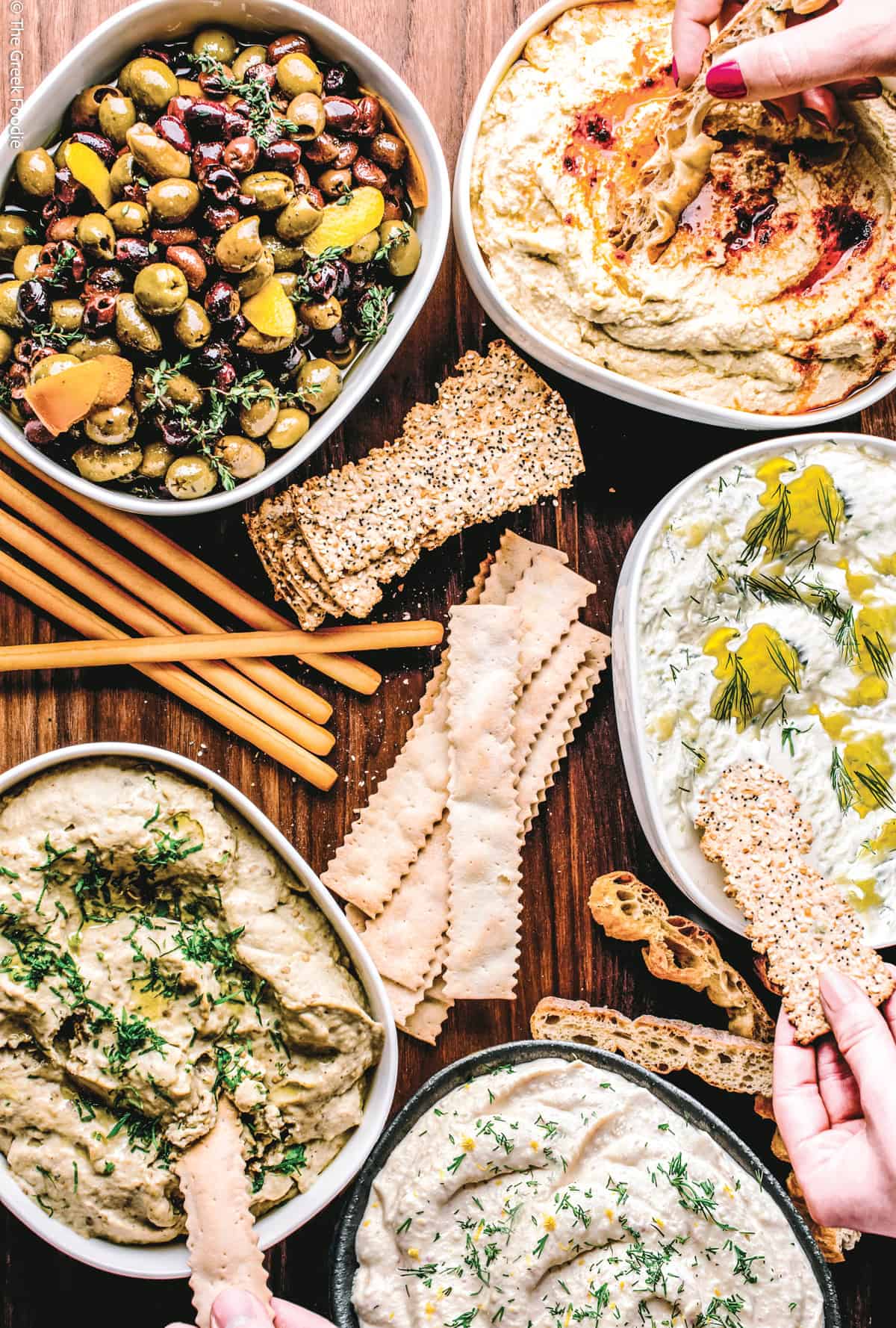 A Greek meze spread with multiple dips including melitzanosalata, tzatziki, and skordalia, served with olives, crackers, and breadsticks on a dark wood table
