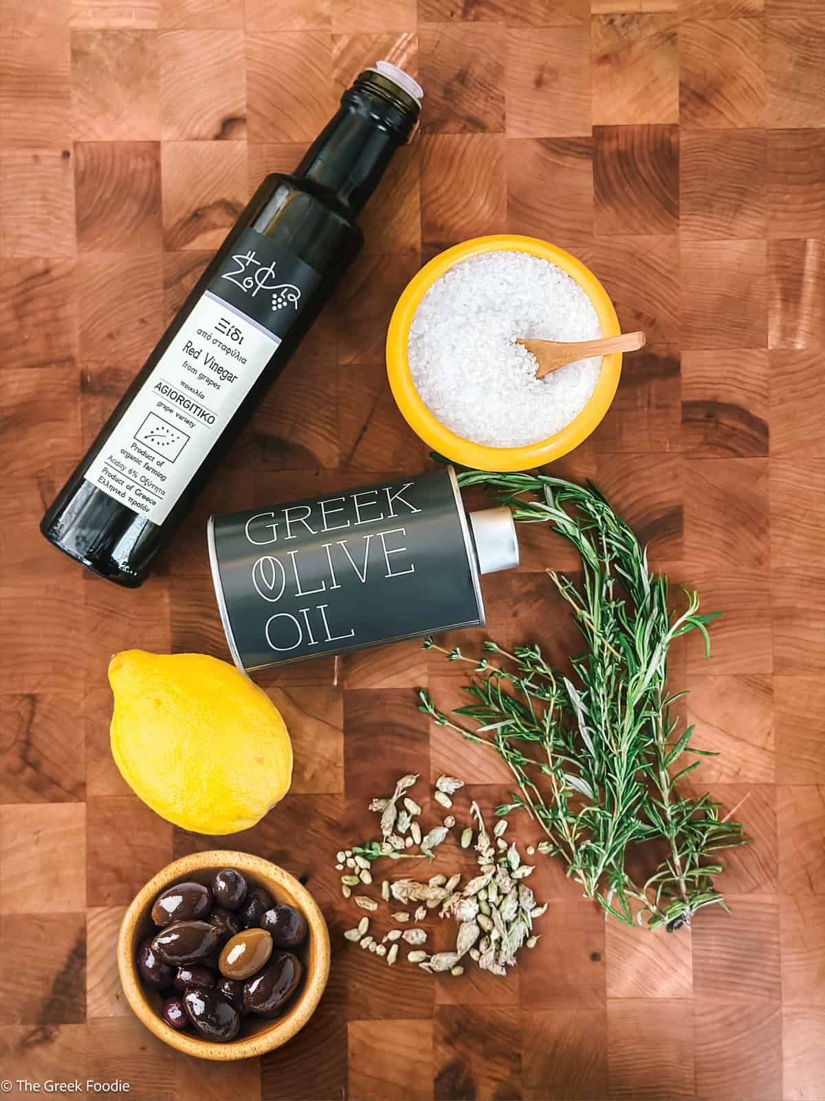 Greek salad dressing ingredients flat lay — Greek olive oil bottle, sea salt, Kalamata olives, lemon, and fresh rosemary on a wooden board
