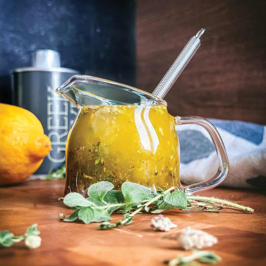 Greek salad dressing in a glass jar with a whisk, fresh oregano, thyme, and lemon on a wooden table