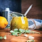 Greek salad dressing in a glass jar with a whisk, fresh oregano, thyme, and lemon on a wooden table