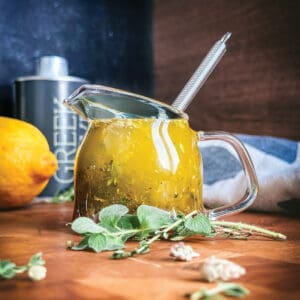 Greek salad dressing in a glass jar with a whisk, fresh oregano, thyme, and lemon on a wooden table