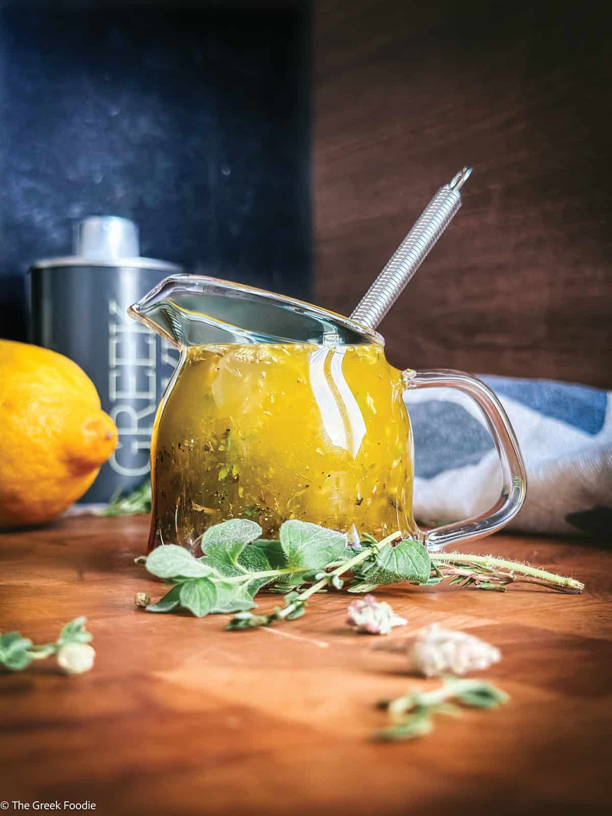 Greek salad dressing in a glass jar with a whisk, fresh oregano, thyme, and lemon on a wooden table