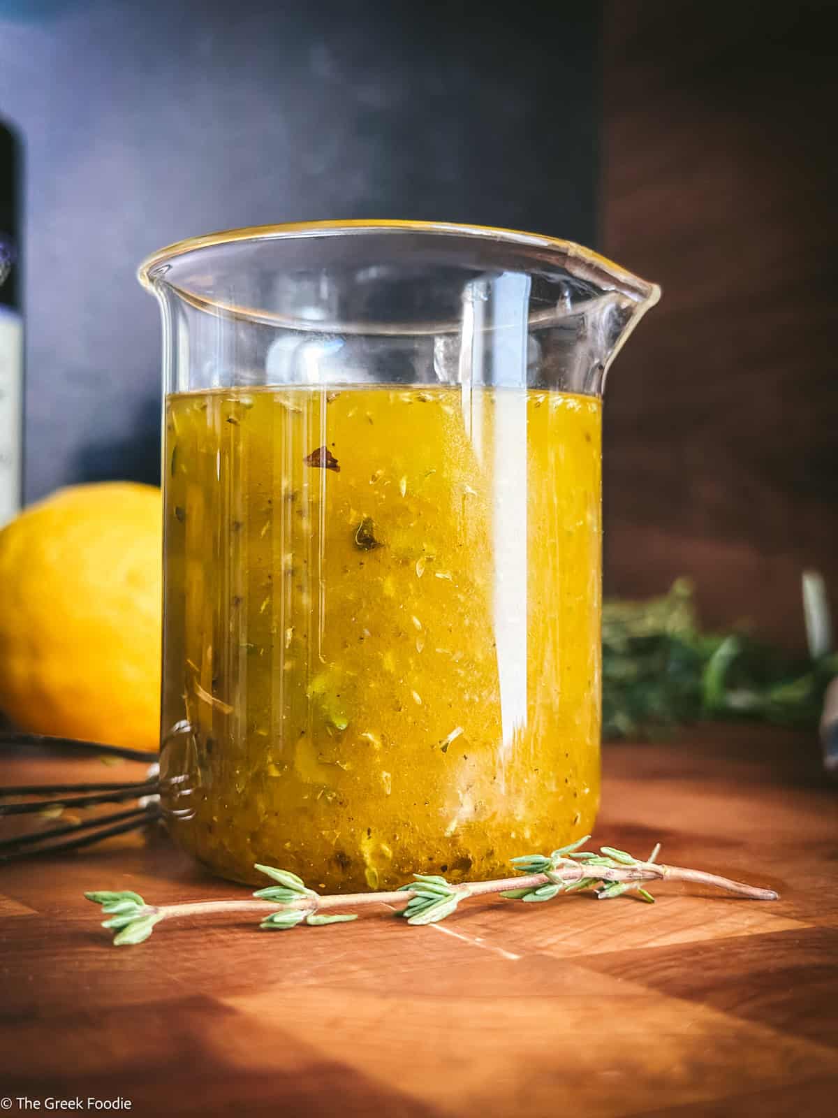 Greek salad dressing in a glass pitcher with fresh oregano and thyme on a wooden surface
