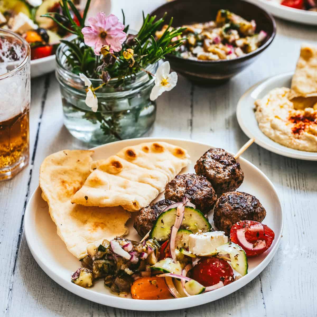 A Greek meze plate with keftedes skewer, Greek salad, melitzanosalata and warm pita bread, surrounded by dips and drinks on a white wooden table