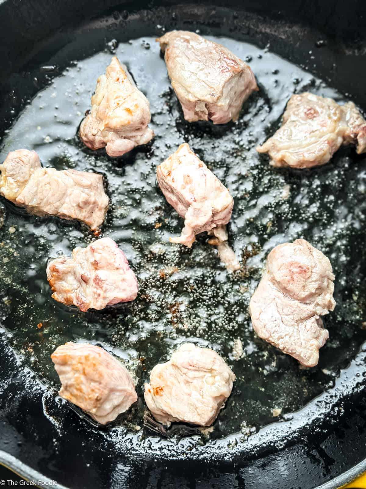 Pork neck pieces searing in a cast iron skillet with olive oil — the first step for bekri meze