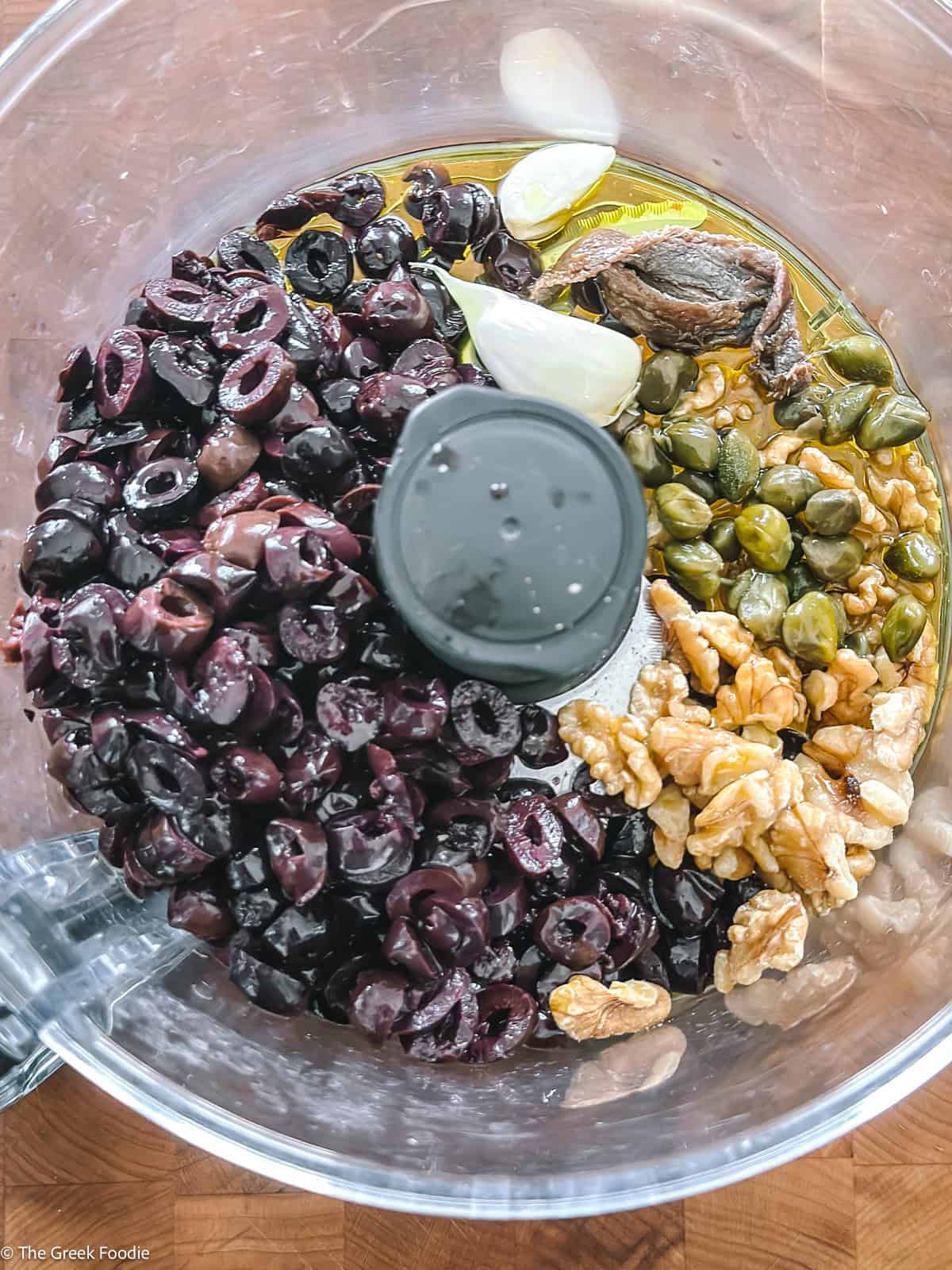 Overhead view of kalamata olive tapenade ingredients in a blender including black Kalamata olives, capers, walnuts, garlic and olive oil