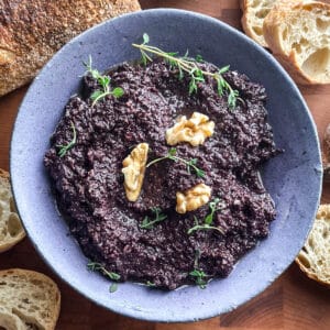 Greek kalamata olive tapenade in a grey bowl topped with a walnut and fresh thyme sprigs, served with toasted bread