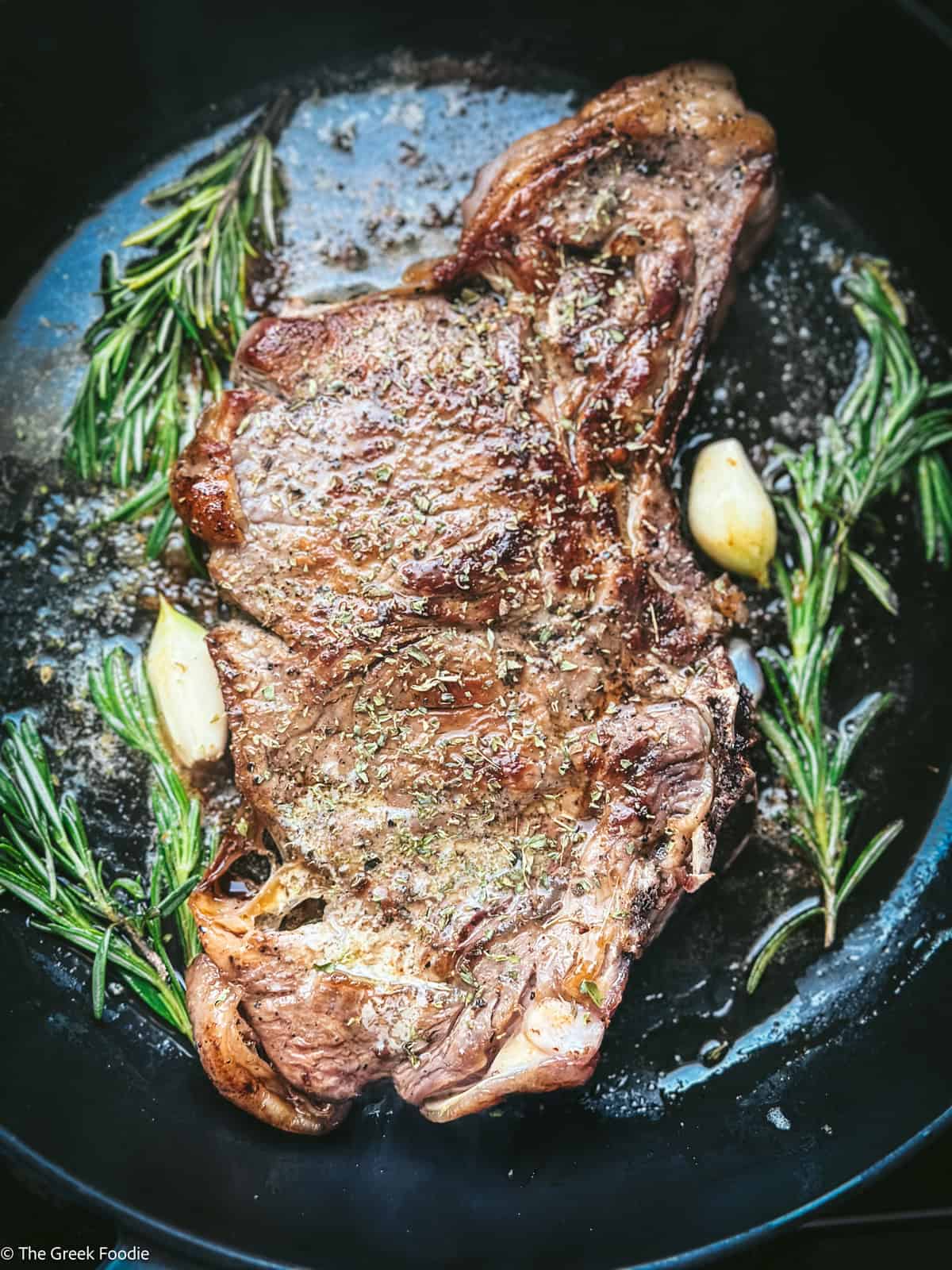 Greek brizola steak in a cast iron pan with a lemon half, rosemary and garlic during the butter baste