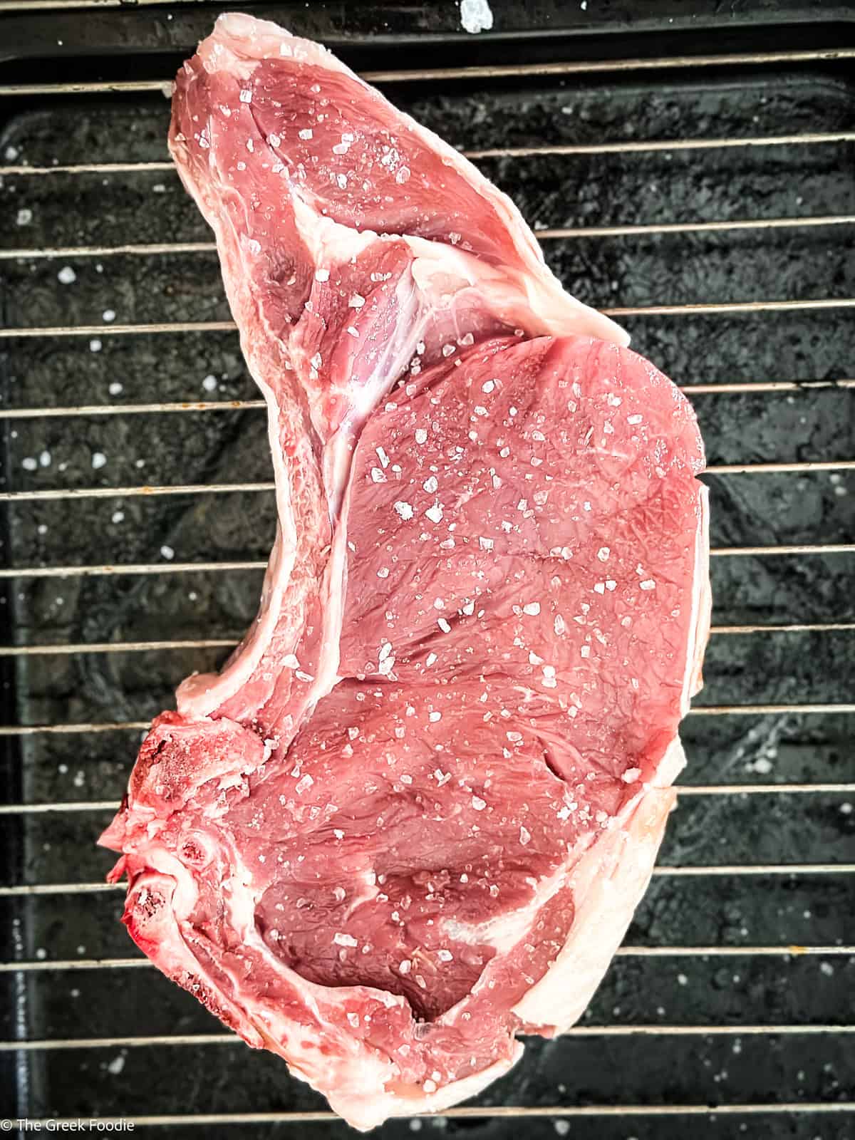 Raw spalobrizola Greek ribeye steak on a rack seasoned generously with coarse salt