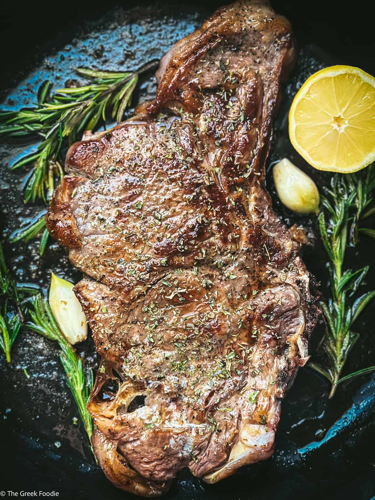 Greek ribeye brizola steak cooked in a cast iron pan with herbs, garlic and lemon wedge