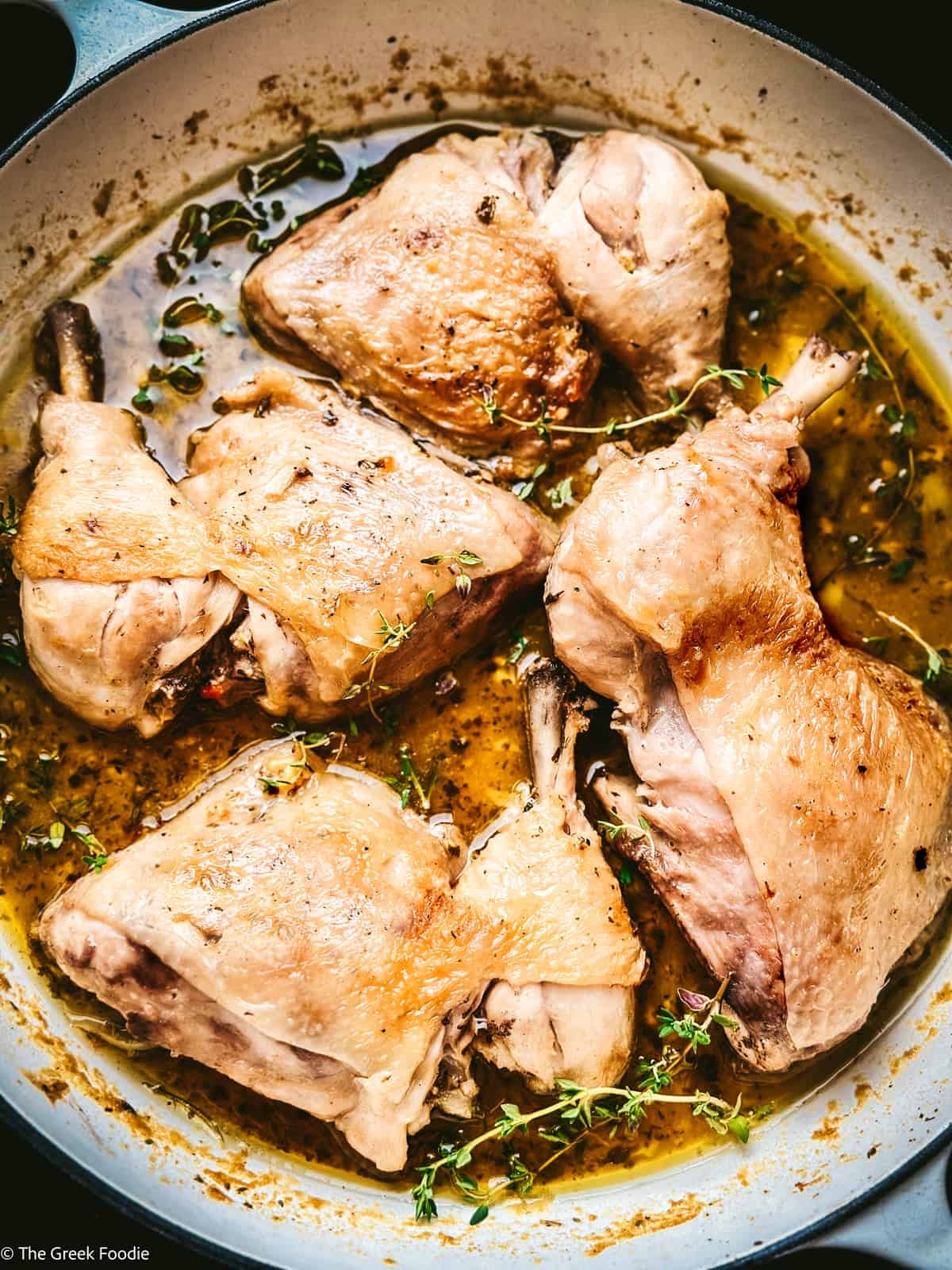 Overhead view of braised chicken thighs and drumsticks in a white Dutch oven with golden lemon herb sauce and fresh thyme sprigs