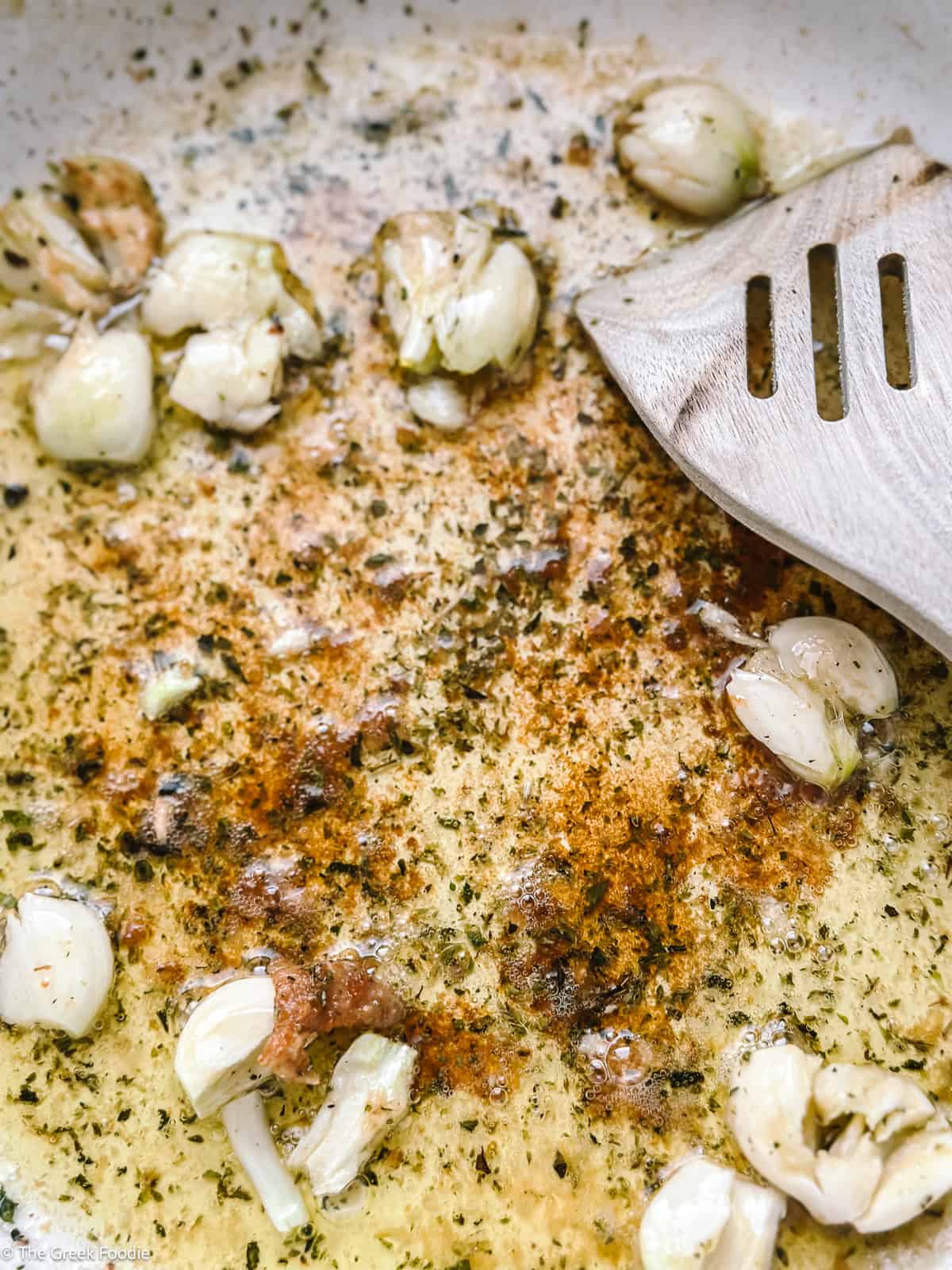 Garlic cloves and dried oregano toasting in olive oil in a white Dutch oven, with a wooden spatula scraping the fond
