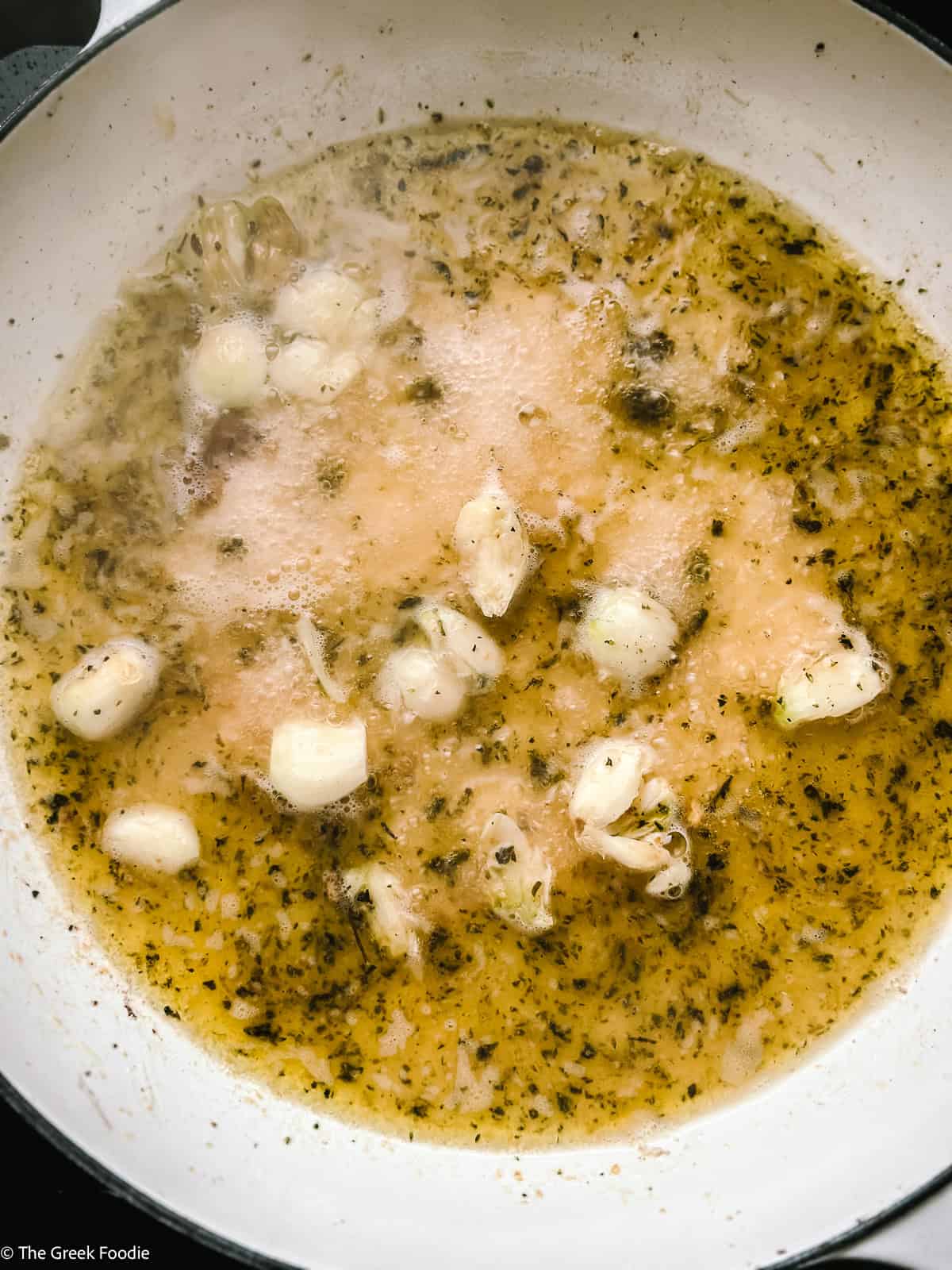 White wine and chicken stock deglazing a white Dutch oven with garlic and herbs, bubbling over medium heat